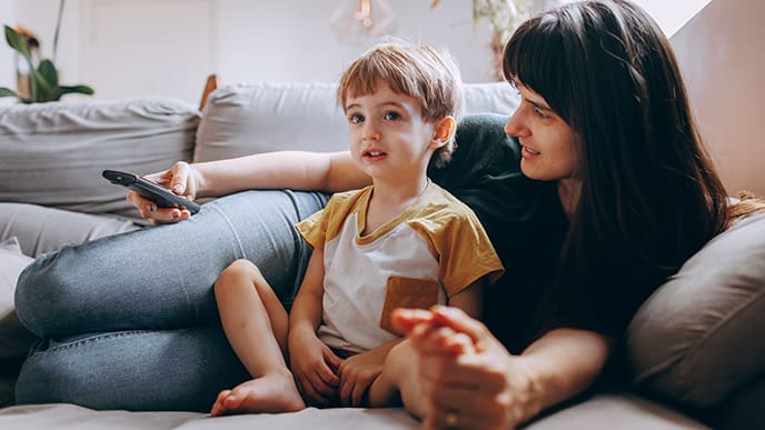 Uma mãe e o seu filho pequeno, deitados no sofá. Ela segura o controle remote da televisão e a criança assiste a um dos canais infantis da TV Desktop.