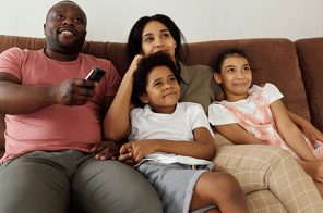 Uma família, composto pelo pai, mãe, um menino e uma menina. Eles estão sentados no sofá e o pai segurando o controle remoto da televisão.