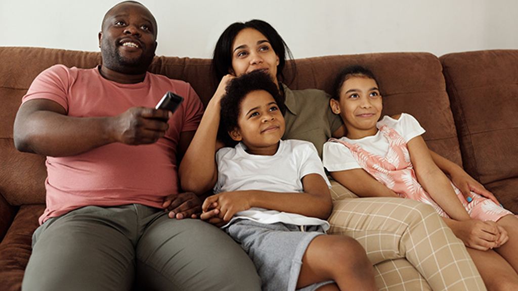 Uma família, composto pelo pai, mãe, um menino e uma menina. Eles estão sentados no sofá e o pai segurando o controle remoto da televisão.