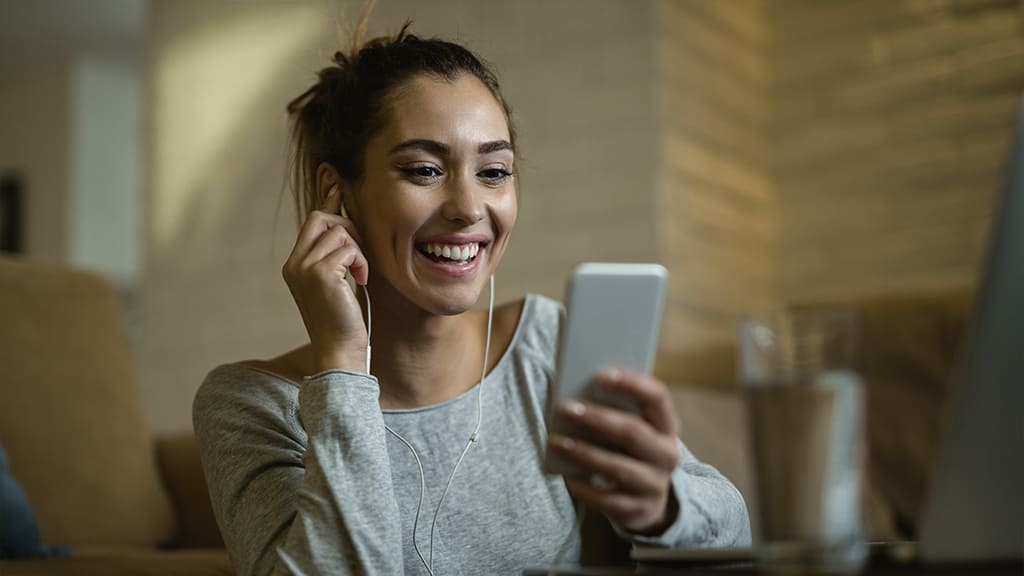 Uma mulher na sala, ela segura um celular e está com fone de ouvido. Ela ouve um audiobook.