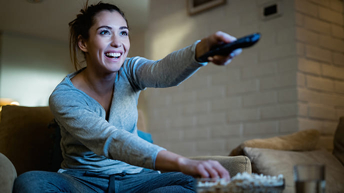 Uma mulher, sentado no sofá, segurando o controle remoto da televisão, mudando o canal da tv por assinatura da Desktop, e comendo pipoca.