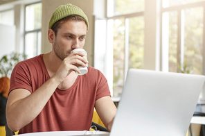 Um homem, em um ambiente interno, sentado à mesa, tomando um café e usando um notebook conectado à internet da Desktop.