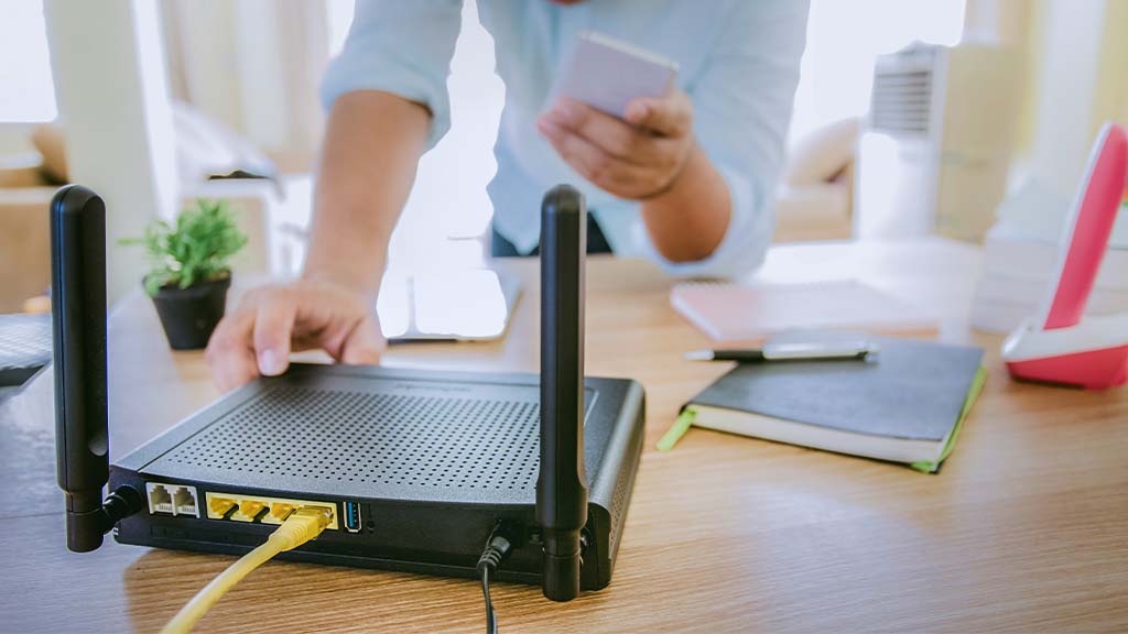 Uma pessoa segurando o celular e ligando o roteador que está sobre uma mesa.