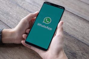 As mãos de uma pessoa posicionada sobre uma mesa de madeira, segurando o celular. Na tela, a imagem principal do app do WhatsApp.
