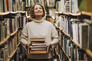 Uma mulher no corredor de uma biblioteca. Ela segura uma pilha de livros.