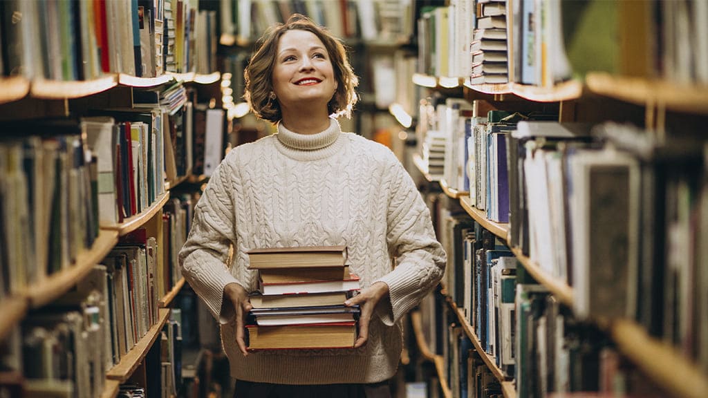 Uma mulher no corredor de uma biblioteca. Ela segura uma pilha de livros.