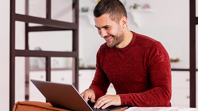 Um homem, usando uma blusa de manga longa, vermelha, na cozinha da casa, usando um notebook conectado à internet da Desktop.