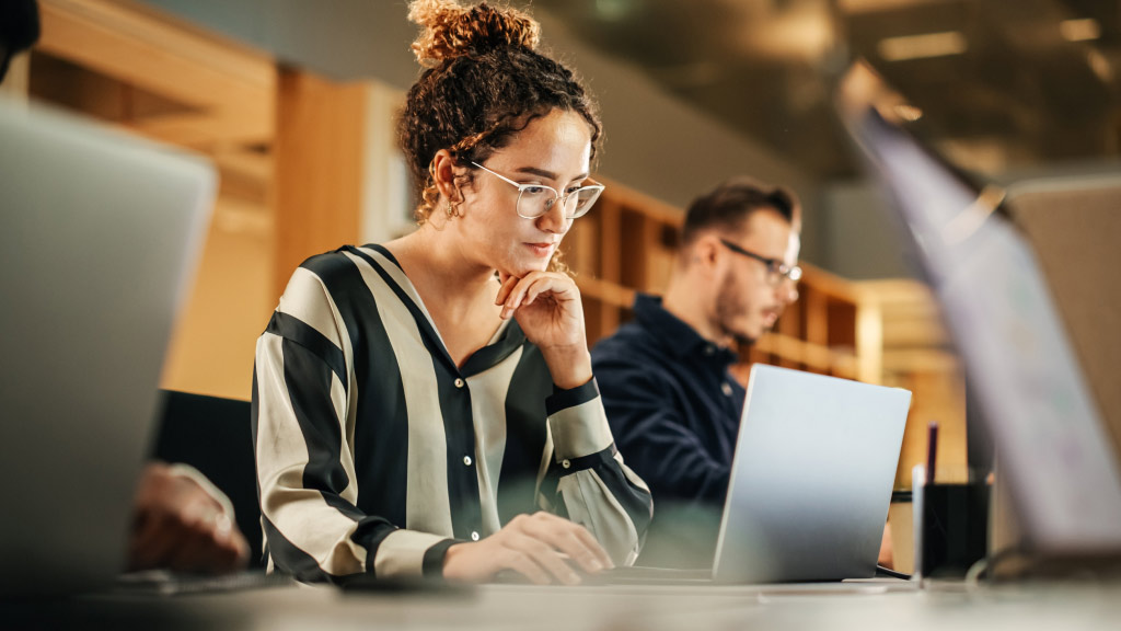 Na imagem, uma mulher trabalhando, ela usa um notebook. Ao fundo um colega de trabalho, também usando um computador.