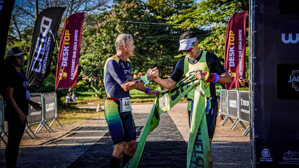 Na imagem, dois atletas se cumprimentam ao final de uma prova de corrida. Ao fundo, dois banners com o logo da Desktop, a internet que joga junto