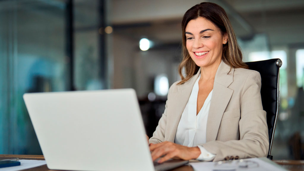 Na imagem, uma mulher, no ambiente de trabalho, com uma camisa branca e um blazer bege, usando o notebook.