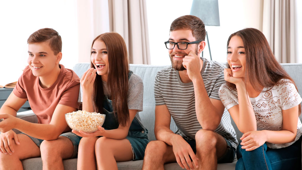 Na imagem, quatro adolescentes sentados no sofá, assistindo TV. Uma das adolescentes segura uma vasilha com pipoca.